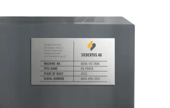 Ecke eines dunkelgrauen Gehäuses mit einem kleinen, natur eloxierten Maschinenschild, das die Aufschrift Kompressor 2 und eine Seriennummer trägt.